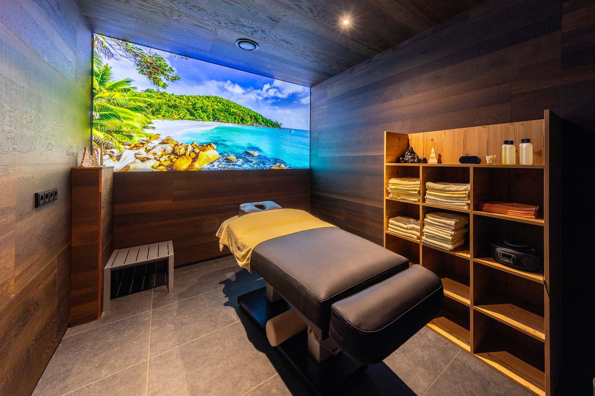 Wellnesshotel in Jena? Moderne Massageraum mit tropischem Strandbild und Holzregal mit Handtüchern
