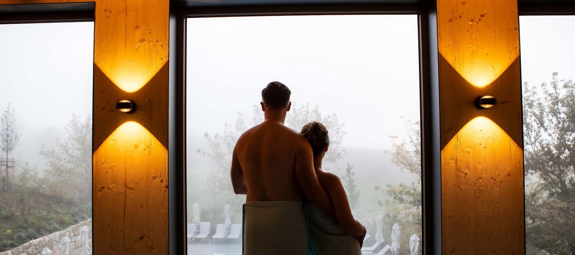 Wellnesshotel in Jena? Paar mit Handtüchern schaut auf Pool und Landschaft durch großes Fenster