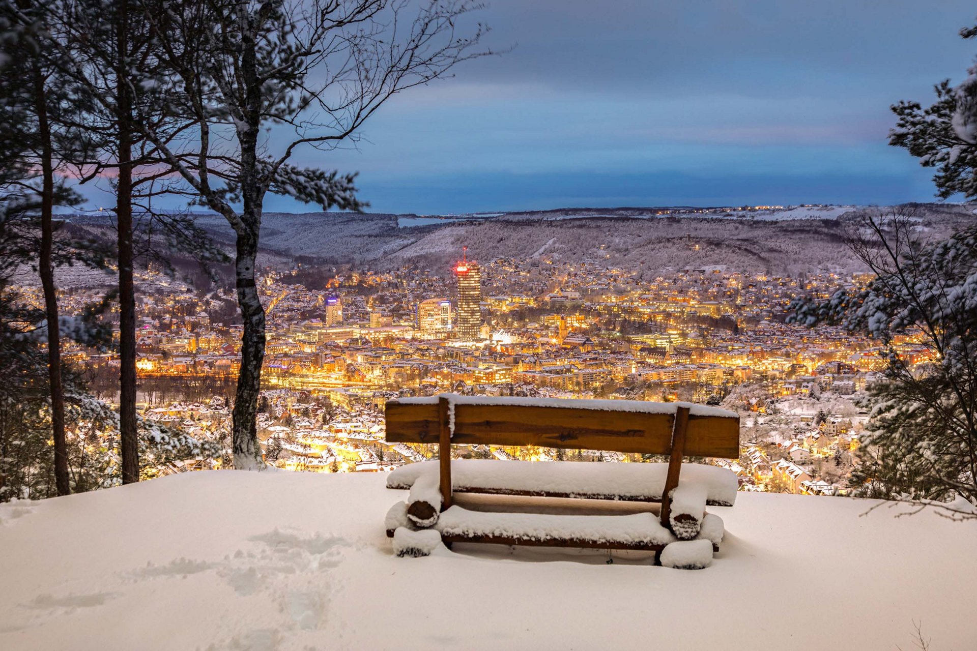 Sehenswürdigkeiten in und um Jena Schneebedehnte Bank mit Blick auf beleuchtete Stadt in einem verschneiten Tal bei Dämmerung