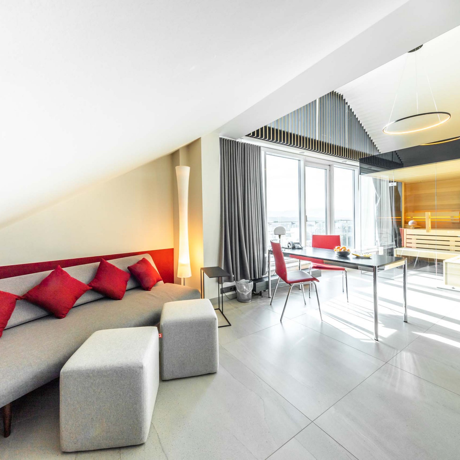 Urlaub in Jena: mein Fair Resort Modernes Wohnzimmer mit grauem Sofa, roten Kissen und Glaswand zur Sauna